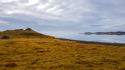 A fjord with a bench on a side. Grass in dried after winter. IN the back invisible line of horizon. Skyline blending with the ocean water. Calmness and serenity. Beauty in the nature.