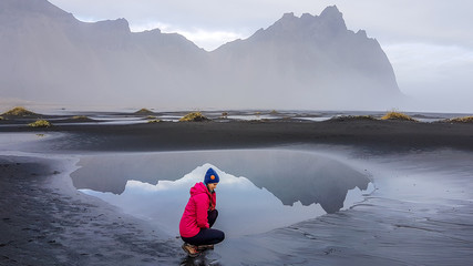 A girl wearing a pink jacket squatting on a black sand beach, in front of a paddle reflecting the mountains in a shallow sea shore. Magical and mysterious hidden gem. Beauty of the nature, Serenity