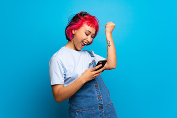 Young woman with pink hair over blue wall with phone in victory position