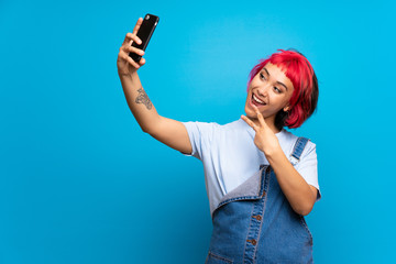 Young woman with pink hair over blue wall making a selfie