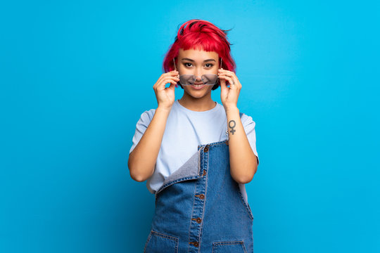 Young Woman With Pink Hair Over Blue Wall With Glasses And Surprised