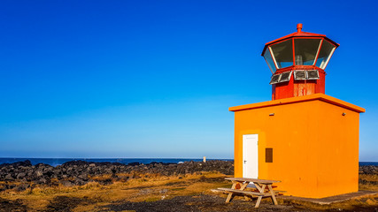 Fototapeta na wymiar A cute little, orange lighthouse placed on the shore of a steep cliff. In front of the lighthouse there is a bench. Orange contrasted with the blue sky. Red top of the tower.