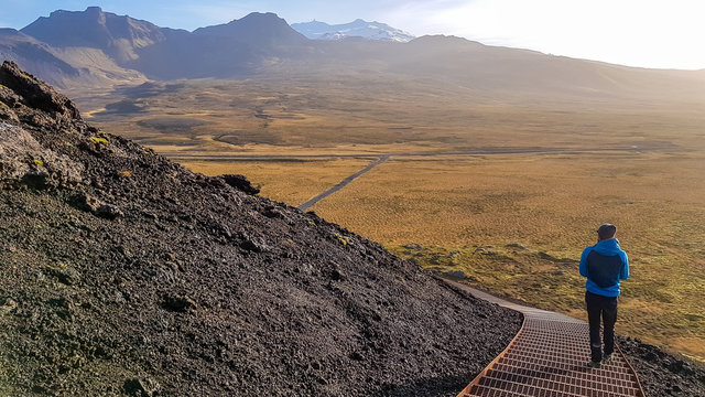 A young man wearing blue jacket walking down the path on the volcano side. Sunny and bright day. Slopes of the volcano overgrown with grass. In the back tall mountains visible. Beauty of the nature