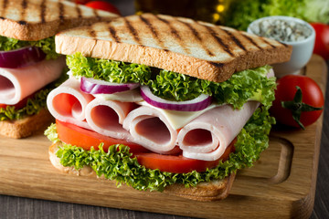 Close-up photo of a club sandwich. Sandwich with meat, prosciutto, salami, salad, vegetables, lettuce, tomato, onion and mustard on a fresh sliced rye bread on wooden background. Olives background.