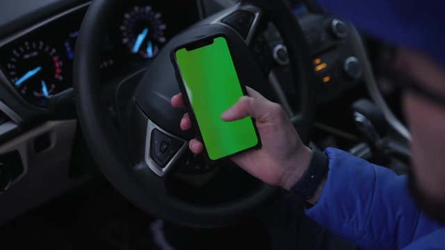 Young man in blue sports clothes sitting at driver's seat in car.  Person using smartphone. Green screen. Touch. Internet, navigator, lifestyle, daily, close-up, indoors