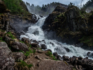 Naklejka na ściany i meble Upper parts of Langfossen waterfall at its splendid. Vast and tall waterfall, flowing around the rocks and in between the trees. Immense power of the nature. The water falls down a towering mountain