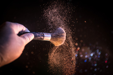 Makeup brush in hand with cosmetic powder on dark background with light and smoke. Powder splash on dark