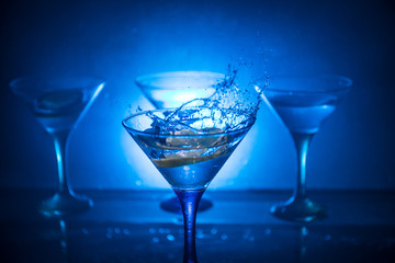 Martini cocktail glass in hand splashing on dark toned smoky background or colorful cocktail in glass with splashes and olives. Party club entertainment. Mixed light. Selective focus