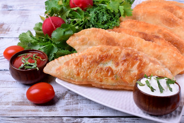 Golden Chebureki on a white plate. Sauces, vegetables and greenery around