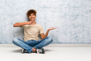 Young african american man sitting on the floor holding copyspace imaginary on the palm to insert...