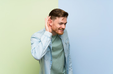 Redhead man over colorful background listening to something by putting hand on the ear