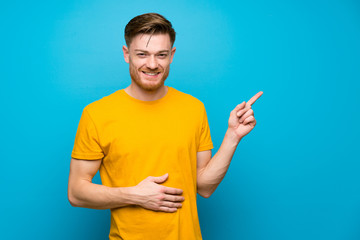 Redhead man over blue wall pointing finger to the side