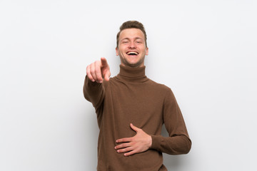 Blonde man over isolated white wall pointing with finger at someone and laughing a lot