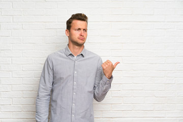 Blonde man over white brick wall unhappy and pointing to the side