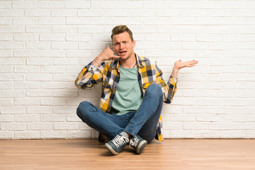 Blonde man sitting on the floor making phone gesture and doubting