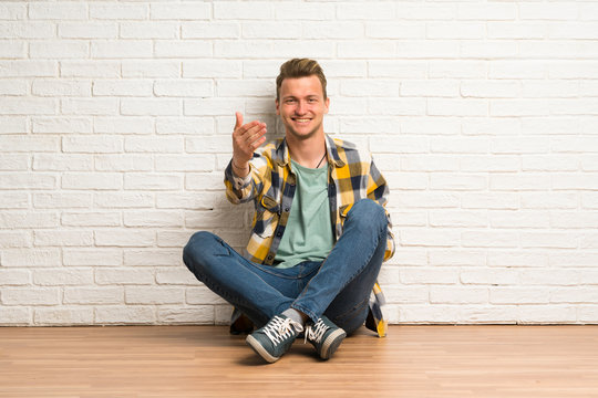 Blonde man sitting on the floor inviting to come with hand. Happy that you came