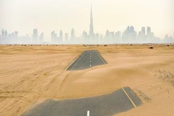 Zelfklevend Fotobehang Luchtfoto van een verlaten weg bedekt met zandduinen in het midden van de woestijn van Dubai. De skyline van Dubai met de Burj Khalifa omgeven door mist op de achtergrond. Dubai, Verenigde Arabische Emiraten. © Travel Wild