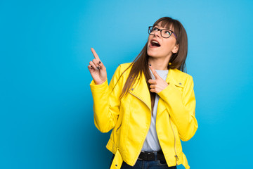 Young woman with yellow jacket on blue background pointing with the index finger and looking up