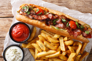 Delicious hot dog with bacon and vegetables served with french fries and sauces close-up. horizontal top view