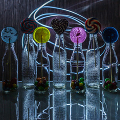 Bright and colorful lollipop and candies in bottles standing on the glass table in the dark on background of lines of light. Concept of fresh drinks. Concept of holidays.