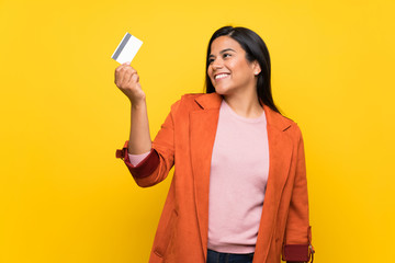 Young Colombian girl over yellow wall holding a credit card and thinking