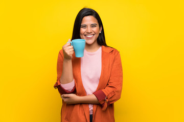 Young Colombian girl over yellow wall holding a hot cup of coffee