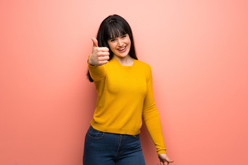 Woman with yellow sweater over pink wall with thumbs up because something good has happened