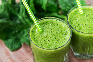 Two green smoothies in glasses with straws, spinach, healthy ingredient on the wooden table