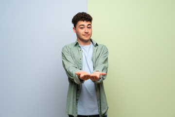 Young man over blue and green background holding copyspace imaginary on the palm to insert an ad