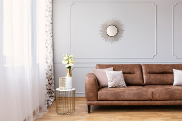 Elegant living room interior with a brown sofa, side table with flowers in a vase and mirror on a wall. Real photo