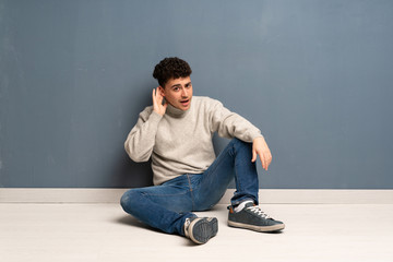 Young man sitting on the floor listening to something by putting hand on the ear