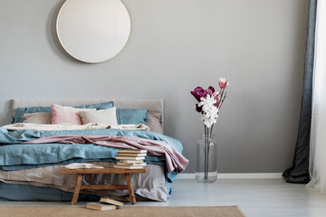 Trendy round wooden mirror on beige wall of classy bedroom interior with large double bed and flowers in vase