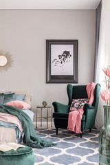 Stylish poster in black frame on grey wall of fashionable bedroom interior with emerald green armchair and king size bed