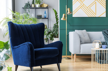 Blue armchair next to grey scandinavian sofa in tropical inspired interior with green and gold colors