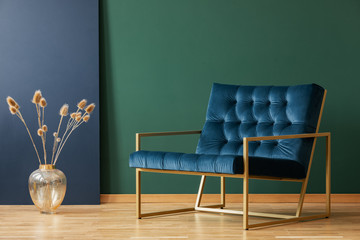 Plant in gold vase next to armchair in blue and green elegant living room interior. Real photo