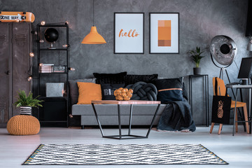 Orange lamp above table and posters above settee in grey living room interior with pouf. Real photo