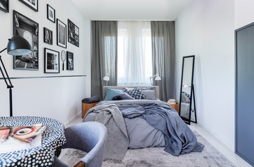 Mirror next to bed with grey sheets in bedroom interior with black lamp and posters. Real photo