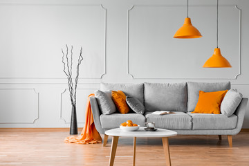 Real photo of simple living room interior with orange lamps, pillows and grey sofa