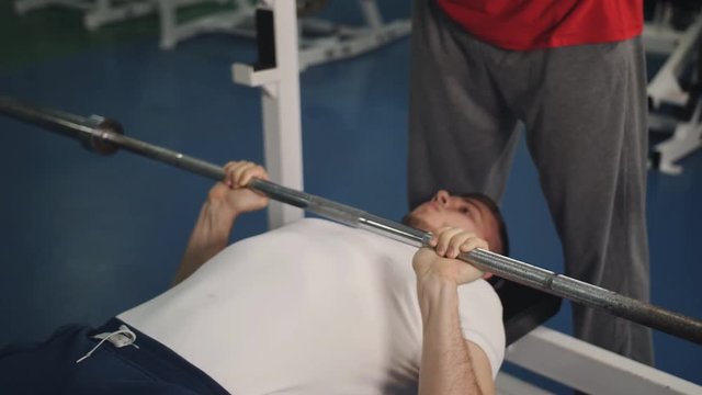 Young athlete is doing bench press with help of trainer in gym. Man raises free weight under control of instructor in sports club. Handsome guy lies on bench and does exercise with iron object