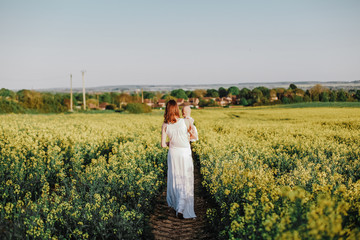 Woman in long white dress walking with baby in the Rape Field. Vilage in background. Rear view.