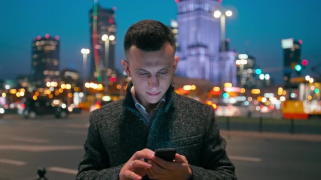 Adult Caucasian Confident Young Business Man Entrepreneur is Using Smartphone App Outdoors in the City in the Late Evening or Night with Beautiful Blurred Cityscape Lights. Medium 4K Static Shot