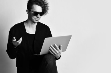 Closeup of young modern man in sunglasses looking at laptop that holds on his knee and  talks about it gesticulating actively.