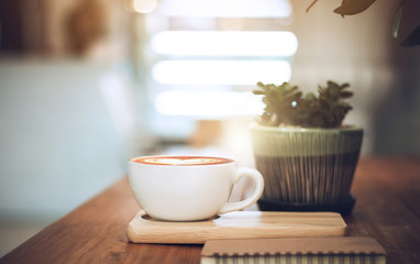 Closeup of latte art coffee on wooden table with book and small tree in ceramic vase background, vintage color tone