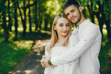 A beautiful and gentle girl with light hair and a white dress is walking in a sunny summer forest with her handsome guy in a white shirt and dark pants