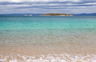 Obraz premium Crystal clear water, rippling gently on the shore gradually turns a pale turquoise and then blue as it reaches the island on the horizon.