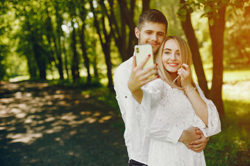 A beautiful and gentle girl with light hair and a white dress is walking in a sunny summer forest with her handsome guy in a white shirt and dark pants and they use the phone