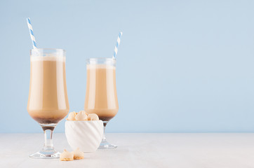 Fresh coffee drinks with cream, striped straws  in wineglass and cookies in bowl on soft light blue wall and white wooden table, copy space.