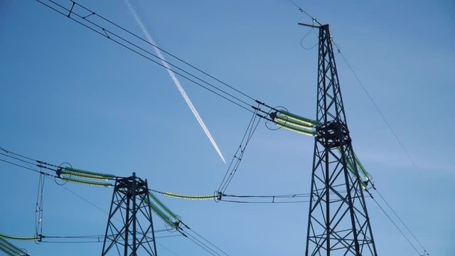 Close up: top of two high voltage power transmission tower and electricity pylon with wires against blue sky. Airplane with white trail on the blue sky