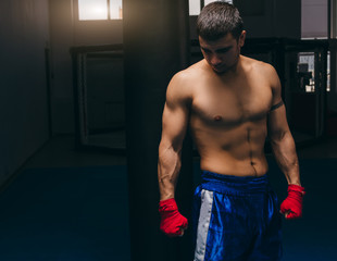 Young caucasian male boxer with muscular bare torso wearing blue boxing shorts and red elastic bandages on hands posing at boxing studio