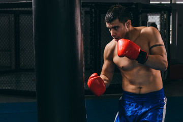 Handsome determinated bare chested male boxer in red gloves getting prepared for big fight, doing cardio boxing workout with punching bag in empty dark gym. Sport, challenge victory, workout.